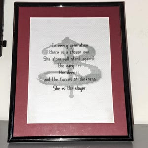 May include: Framed cross-stitch art with a black frame and a burgundy mat. The white fabric features a quote: "In every generation there is a chosen one. She is the slayer."
