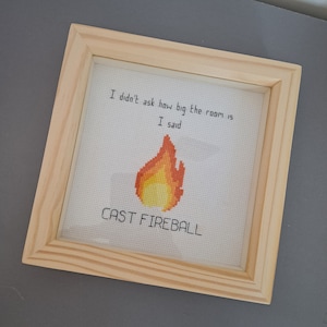May include: A framed cross-stitch with a light brown frame. The cross-stitch features a yellow and orange pixelated flame with the text "I didn't ask how big the room is I said CAST FIREBALL".
