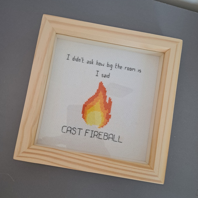 May include: A framed cross-stitch with a light brown frame. The cross-stitch features a yellow and orange pixelated flame with the text "I didn't ask how big the room is I said CAST FIREBALL".
