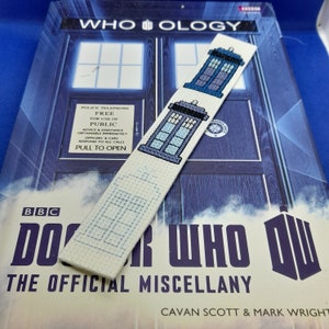 May include: A white fabric bookmark with a blue and white embroidered image of the TARDIS, the time machine from the Doctor Who television series. The bookmark is lying on top of a Doctor Who book.