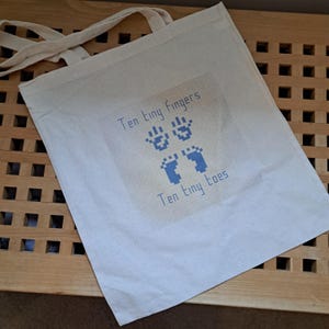 May include: A white tote bag with a blue design of hands and feet. The text "Ten tiny fingers" and "Ten tiny toes" is printed on the bag.