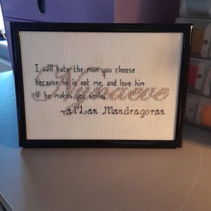 May include: A framed cross-stitch piece on white Aida cloth with black text that reads: I will hate the man you choose because he is not me, and love him if he makes you smile. ~al'Lan Mandragoran. The frame is a simple, glossy black rectangular border.