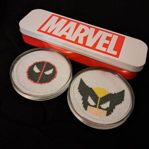 May include: Two round white coasters with red and black embroidered images of Marvel characters Deadpool and Wolverine. The coasters are in front of a red and white tin box with the word "MARVEL" printed in red.