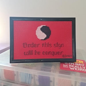 May include: A framed cross-stitch piece on a red background featuring a white and black yin-yang symbol. The black text reads "Under this sign will he conquer" with a copyright symbol and the year 2023 in the bottom right corner.