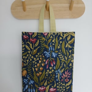 May include: A tote bag featuring a floral design in navy, red, and yellow. The bag has a pale yellow handle and is suspended from a wooden wall hook with three pegs.