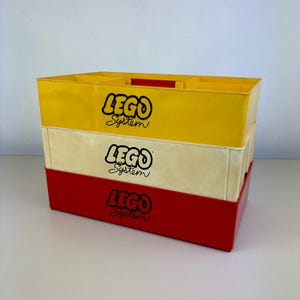 May include: Three stacked vintage LEGO System storage boxes. The top box is yellow, the middle is off-white, and the bottom is red. Each box has the LEGO System logo in black.