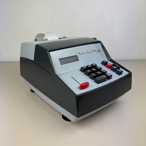 Op de afbeelding: Een vintage Remington 44 rekenmachine in grijs en zwart met rode en zwarte toetsen. De rekenmachine heeft een papieren rol en een displayvenster. De rekenmachine staat op een lichtbeige oppervlak.