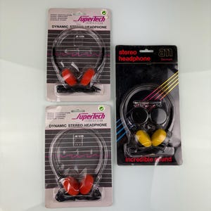 Puede incluir: Tres auriculares estéreo vintage en su embalaje original. Dos son de la marca "Supertech" con almohadillas rojas, y uno es de la marca "am" con almohadillas amarillas. El embalaje incluye el texto "DYNAMIC STEREO HEADPHONE".