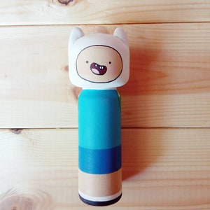 Puede incluir: Una mu&ntilde;eca de madera de Finn el Humano de la serie animada Adventure Time. Finn lleva una camisa azul, pantalones cortos azules y un sombrero blanco con una cara sonriente.