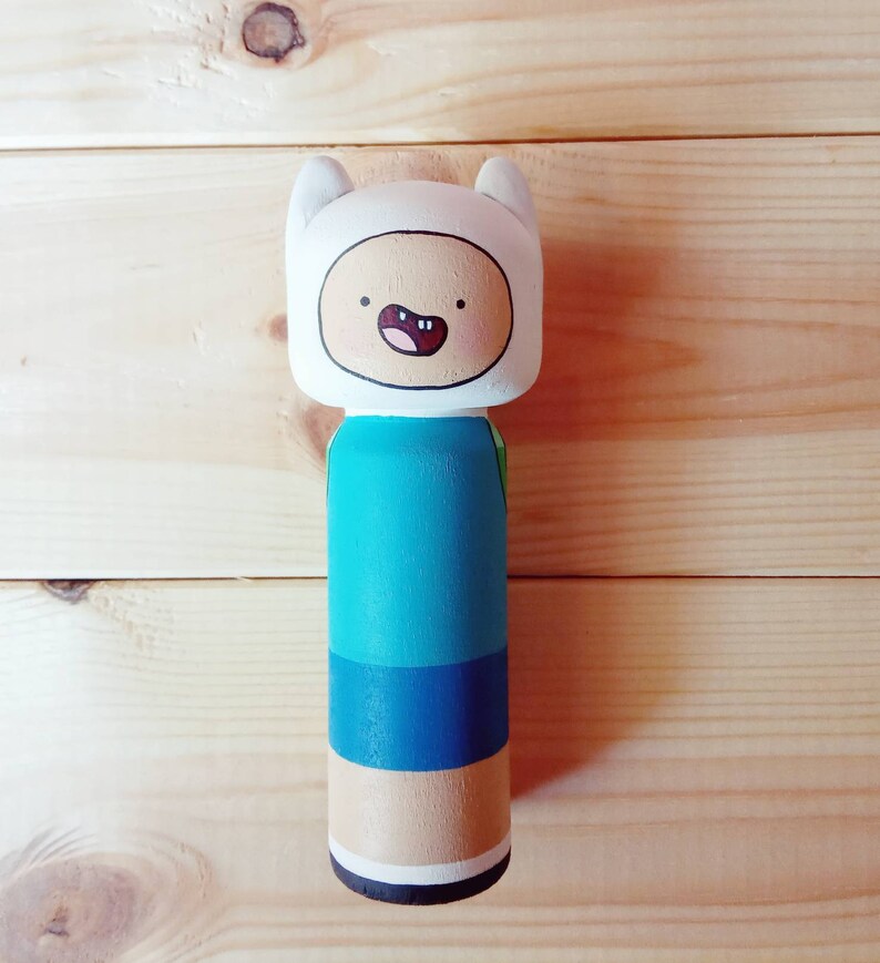Puede incluir: Una mu&ntilde;eca de madera de Finn el Humano de la serie animada Adventure Time. Finn lleva una camisa azul, pantalones cortos azules y un sombrero blanco con una cara sonriente.