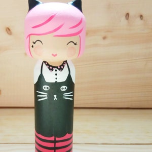 Puede incluir: Una mu&ntilde;eca de madera pintada a mano con cabello rosado, orejas de gato negras y un vestido negro con cuello blanco y una cara de gato negro en la parte delantera. La mu&ntilde;eca lleva leggings a rayas rojas y negras.