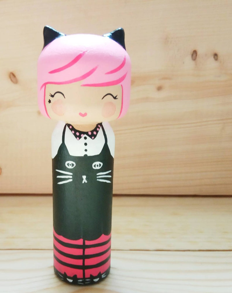 Puede incluir: Una mu&ntilde;eca de madera pintada a mano con cabello rosado, orejas de gato negras y un vestido negro con cuello blanco y una cara de gato negro en la parte delantera. La mu&ntilde;eca lleva leggings a rayas rojas y negras.