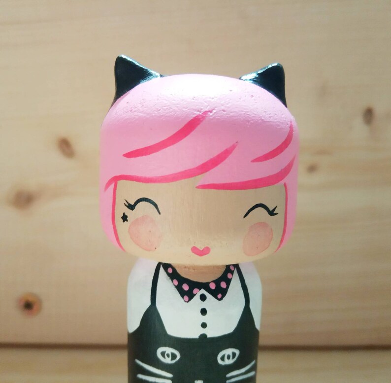 Puede incluir: Una mu&ntilde;eca de madera pintada a mano con cabello rosa, orejas de gato negras y un atuendo blanco y negro. La mu&ntilde;eca tiene una expresi&oacute;n dulce con los ojos cerrados y una peque&ntilde;a boca en forma de coraz&oacute;n.