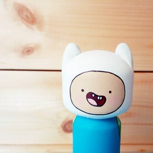 Puede incluir: Una figurita de madera pintada a mano de Finn el Humano de la serie animada Adventure Time. Finn lleva su atuendo azul y verde caracter&iacute;stico y tiene un sombrero blanco con una cara sonriente.