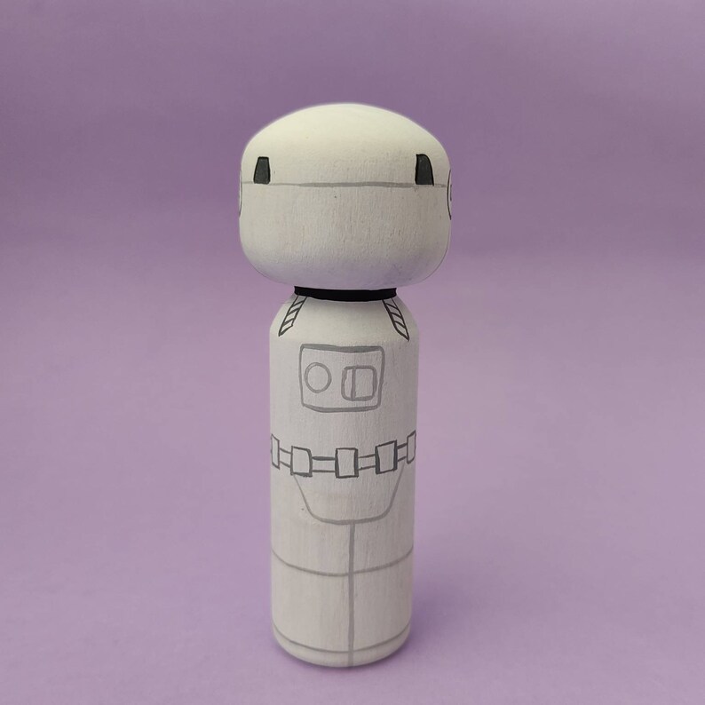 May include: A white wooden doll with black details, resembling a robot or astronaut. The doll has a round head with two black circles for eyes and a black line for a mouth. The body is painted white with black details, including a belt with rectangular buckles and a rectangular panel on the chest with two circles.
