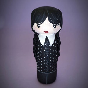 Puede incluir: Una mu&ntilde;eca de madera pintada a mano con cabello negro, una camisa blanca con cuello y lunares negros y una falda negra. La mu&ntilde;eca tiene una cara pintada con ojos negros, labios rojos y mejillas sonrosadas.