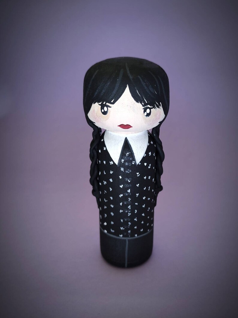 Puede incluir: Una mu&ntilde;eca de madera pintada a mano con cabello negro, una camisa blanca con cuello y lunares negros y una falda negra. La mu&ntilde;eca tiene una cara pintada con ojos negros, labios rojos y mejillas sonrosadas.