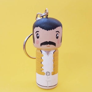 Puede incluir: Un llavero de madera con una ilustraci&oacute;n de dibujos animados de una persona con cabello negro, bigote y una chaqueta amarilla. La persona lleva una camisa blanca y pantalones blancos. El llavero tiene un anillo plateado.