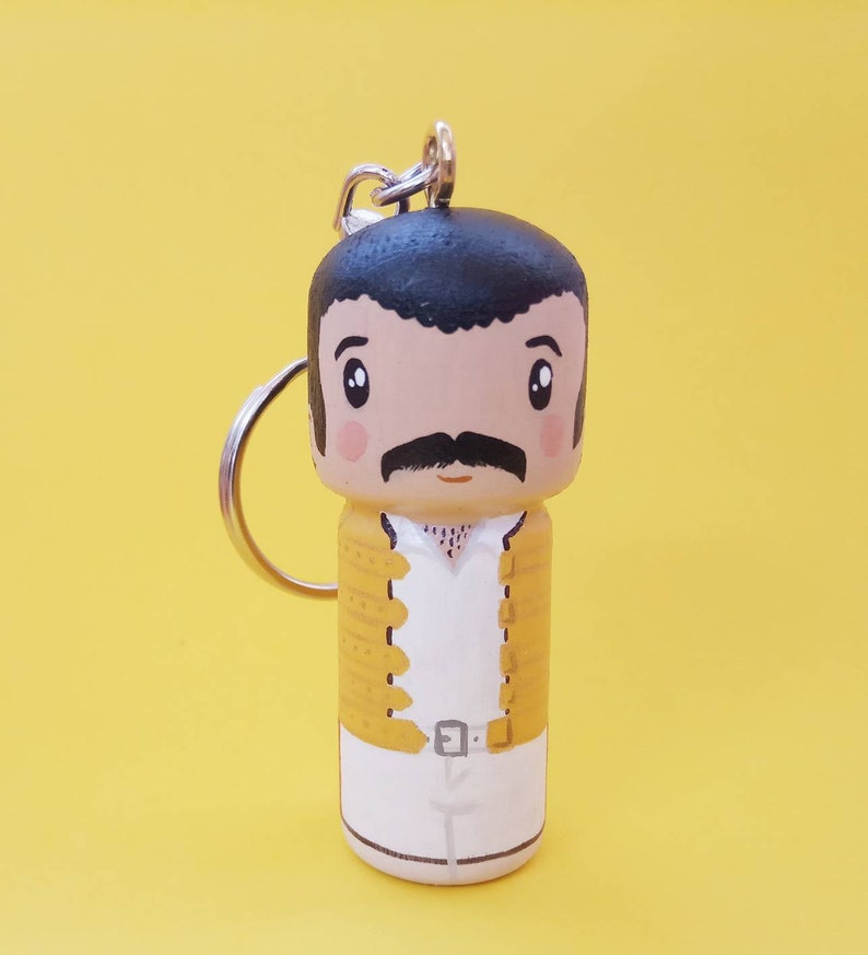 Puede incluir: Un llavero de madera con una ilustraci&oacute;n de dibujos animados de una persona con cabello negro, bigote y una chaqueta amarilla. La persona lleva una camisa blanca y pantalones blancos. El llavero tiene un anillo plateado.