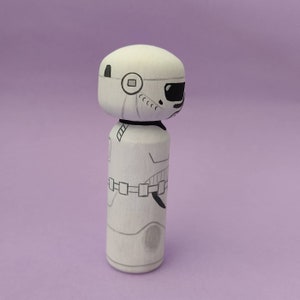 May include: A white wooden doll with a black and white painted design of a Stormtrooper from Star Wars. The doll is standing on a purple background.