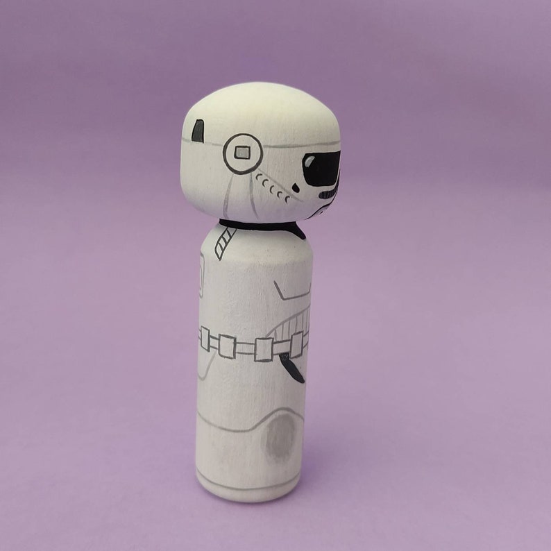 May include: A white wooden doll with a black and white painted design of a Stormtrooper from Star Wars. The doll is standing on a purple background.