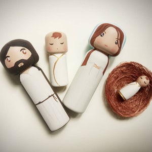 Puede incluir: Un conjunto de cuatro figuras de madera que representan a la Sagrada Familia. Las figuras est&aacute;n pintadas de blanco con cabello y rasgos faciales marrones. El beb&eacute; Jes&uacute;s est&aacute; acostado en un nido marr&oacute;n.