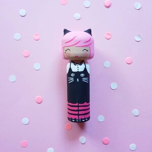 Puede incluir: Una mu&ntilde;eca de madera rosa y negra con un dise&ntilde;o de gato negro en la parte delantera. La mu&ntilde;eca tiene el pelo rosa, los ojos negros y un cuello blanco. La mu&ntilde;eca est&aacute; de pie sobre un fondo rosa con confeti blanco y rosa.