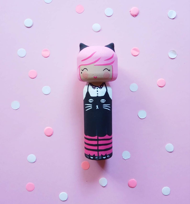 Puede incluir: Una mu&ntilde;eca de madera rosa y negra con un dise&ntilde;o de gato negro en la parte delantera. La mu&ntilde;eca tiene el pelo rosa, los ojos negros y un cuello blanco. La mu&ntilde;eca est&aacute; de pie sobre un fondo rosa con confeti blanco y rosa.