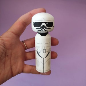 May include: A white wooden doll painted to look like a Stormtrooper from Star Wars. The doll has a black visor and a black belt with white details.