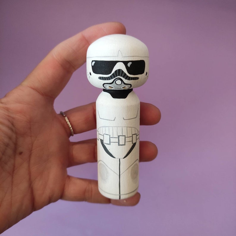 May include: A white wooden doll painted to look like a Stormtrooper from Star Wars. The doll has a black visor and a black belt with white details.