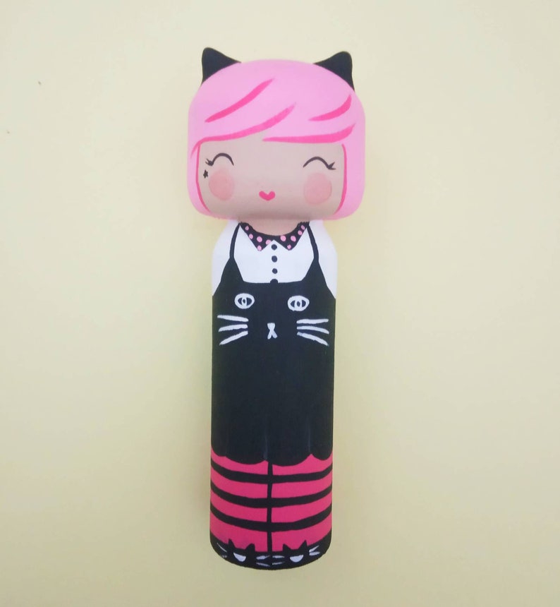 Puede incluir: Una mu&ntilde;eca de madera pintada a mano con pelo rosa, orejas de gato negras y un vestido negro con una cara de gato blanca. La mu&ntilde;eca lleva leggings a rayas rosa y negro y zapatos negros.