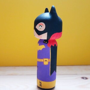 K&ouml;nnte beinhalten: Eine handbemalte Holzpuppe eines Superhelden mit einem schwarzen Umhang, einem violetten Anzug, einem gelben G&uuml;rtel und gelben Stiefeln. Die Puppe tr&auml;gt eine schwarze Maske mit einem roten Akzent und ein gelbes Symbol auf der Brust.