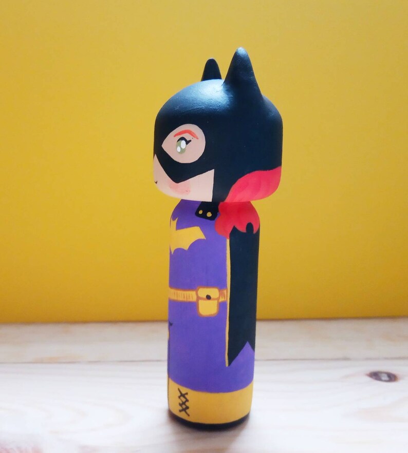 K&ouml;nnte beinhalten: Eine handbemalte Holzpuppe eines Superhelden mit einem schwarzen Umhang, einem violetten Anzug, einem gelben G&uuml;rtel und gelben Stiefeln. Die Puppe tr&auml;gt eine schwarze Maske mit einem roten Akzent und ein gelbes Symbol auf der Brust.