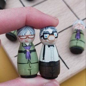 May include: Two hand-painted wooden figurines of an elderly couple. The woman is wearing a green dress with a purple scarf and glasses. The man is wearing a white shirt with a black bow tie, brown trousers, and glasses.