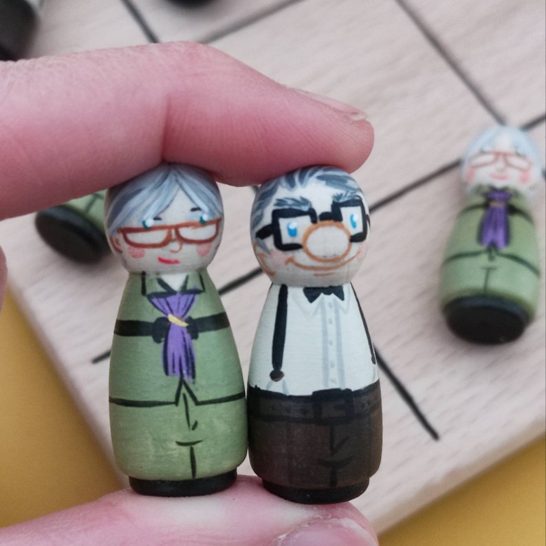 May include: Two hand-painted wooden figurines of an elderly couple. The woman is wearing a green dress with a purple scarf and glasses. The man is wearing a white shirt with a black bow tie, brown trousers, and glasses.