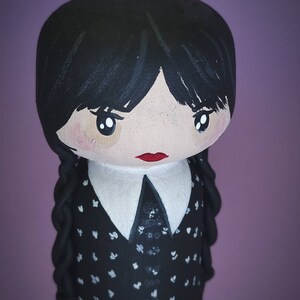 Puede incluir: Una mu&ntilde;eca de madera pintada a mano con cabello negro, un cuello blanco y un vestido negro con lunares blancos. La mu&ntilde;eca tiene una boca roja y ojos marrones grandes.