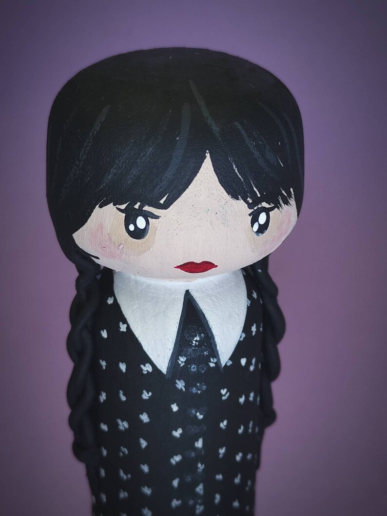 Puede incluir: Una mu&ntilde;eca de madera pintada a mano con cabello negro, un cuello blanco y un vestido negro con lunares blancos. La mu&ntilde;eca tiene una boca roja y ojos marrones grandes.