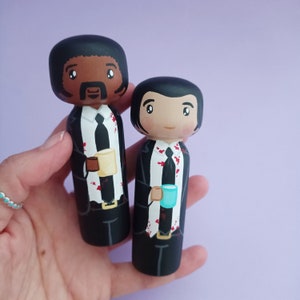 May include: Two wooden peg dolls dressed as characters from the film Pulp Fiction. The dolls are holding coffee cups and are painted in black, white and red.