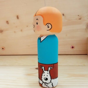 May include: A wooden figurine of a cartoon character with orange hair, a blue shirt, and brown pants. The character is holding a white dog with black spots.
