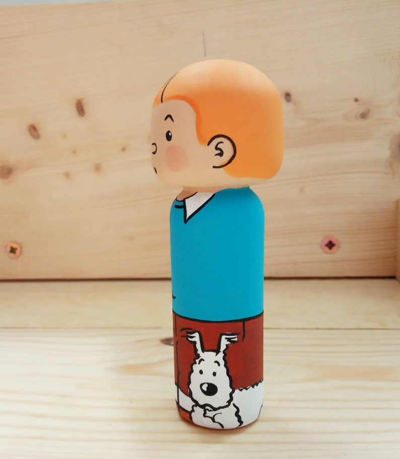 May include: A wooden figurine of a cartoon character with orange hair, a blue shirt, and brown pants. The character is holding a white dog with black spots.