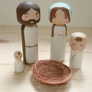 Puede incluir: Conjunto de figuras de madera de la Sagrada Familia. Jos&eacute;, Mar&iacute;a y el ni&ntilde;o Jes&uacute;s est&aacute;n pintados en un estilo de dibujos animados. El ni&ntilde;o Jes&uacute;s est&aacute; en un nido tejido marr&oacute;n.