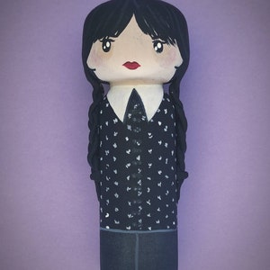 Puede incluir: Una mu&ntilde;eca de madera pintada a mano con el pelo negro en trenzas, que lleva una camisa de lunares negros y blancos, una falda negra y un cuello blanco.