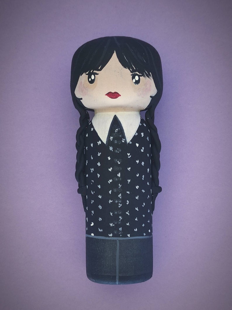 Puede incluir: Una mu&ntilde;eca de madera pintada a mano con el pelo negro en trenzas, que lleva una camisa de lunares negros y blancos, una falda negra y un cuello blanco.