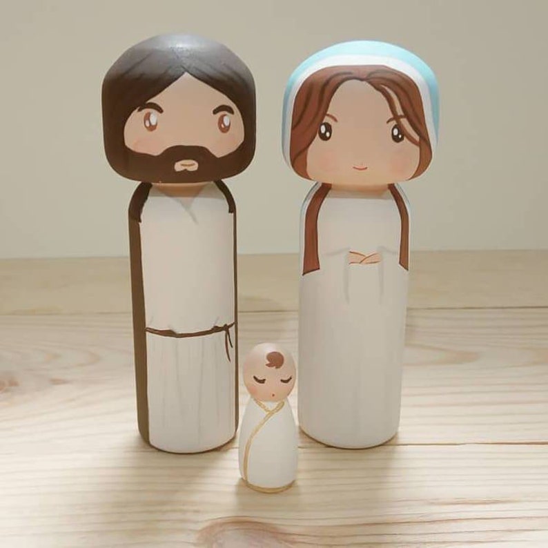 Puede incluir: Un conjunto de tres mu&ntilde;ecos de madera que representan a la Sagrada Familia. Las figuras est&aacute;n pintadas en un estilo de dibujos animados con cabello casta&ntilde;o, ojos marrones y ropa sencilla. La figura del padre lleva una t&uacute;nica marr&oacute;n, la figura de la madre lleva una t&uacute;nica blanca con un velo azul, y la figura del beb&eacute; lleva una t&uacute;nica blanca con un halo dorado.