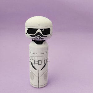 May include: A white painted wooden doll with a black and white stormtrooper design. The doll has a black visor and a black and white design on the body.