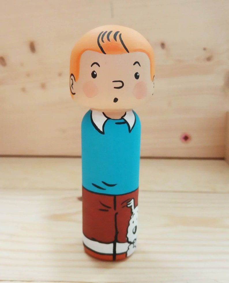 May include: A wooden peg doll painted to look like a cartoon character. The doll has orange hair, a blue shirt, and brown pants. The doll is holding a white dog.