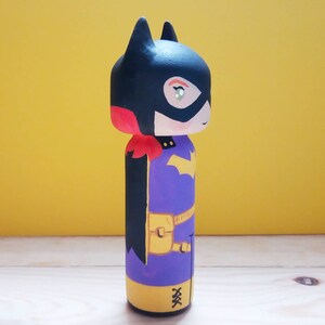 K&ouml;nnte beinhalten: Eine handbemalte Holzpuppe von Batgirl, einer Superheldin aus DC Comics. Die Puppe ist in Schwarz, Rot, Gelb und Lila bemalt und hat einen schwarzen Umhang und einen gelben G&uuml;rtel.