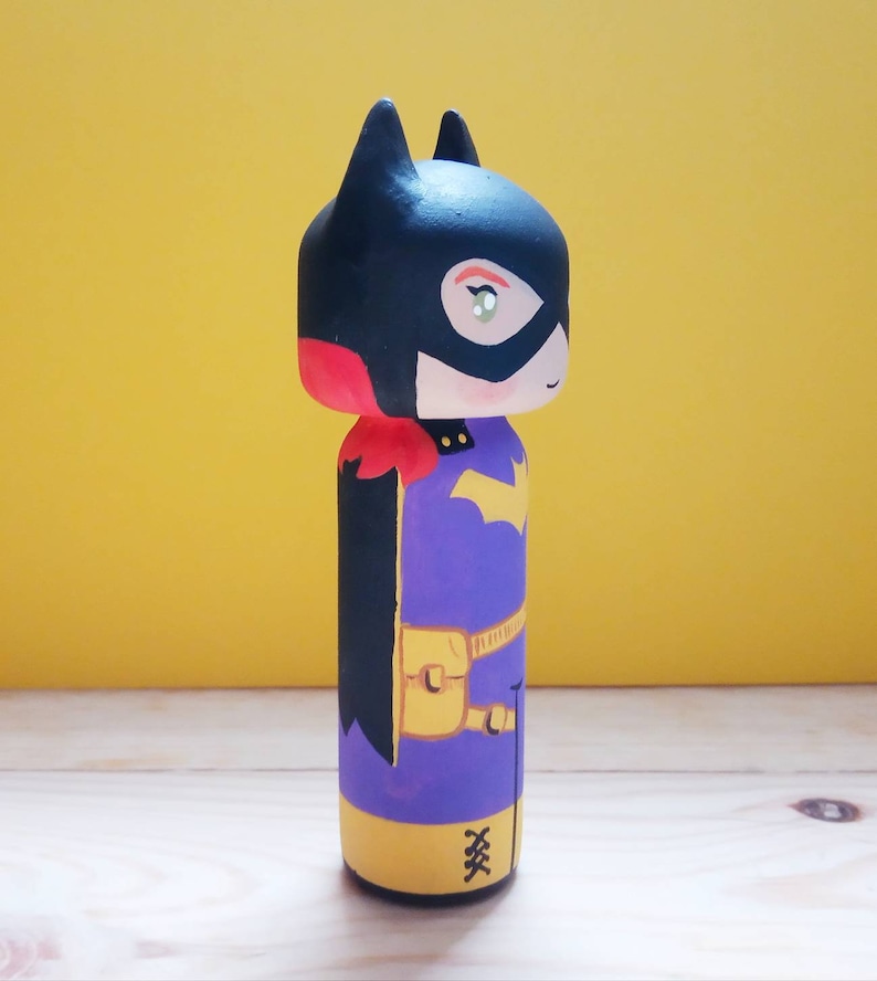 K&ouml;nnte beinhalten: Eine handbemalte Holzpuppe von Batgirl, einer Superheldin aus DC Comics. Die Puppe ist in Schwarz, Rot, Gelb und Lila bemalt und hat einen schwarzen Umhang und einen gelben G&uuml;rtel.