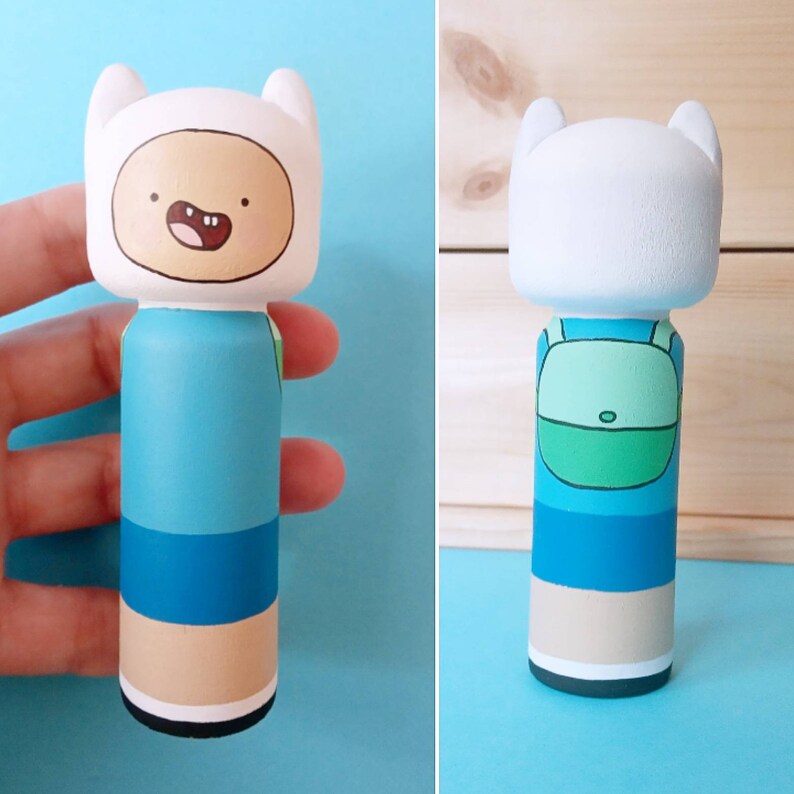 Puede incluir: Una mu&ntilde;eca de madera pintada a mano de Finn el Humano de la serie animada Adventure Time. La mu&ntilde;eca lleva un atuendo azul y verde con un sombrero blanco y una cara sonriente.