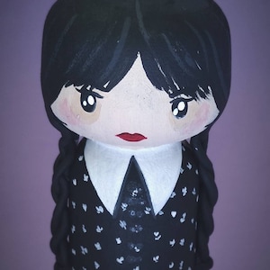 Puede incluir: Una mu&ntilde;eca de madera pintada a mano con cabello negro, un cuello blanco y un vestido negro con lunares blancos. La mu&ntilde;eca tiene una cara pintada con ojos grandes, labios rojos y mejillas sonrosadas.